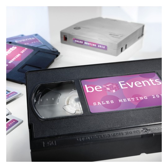 Изображение содержит видеокассету VHS с надписью "beEvents SALES MEETING 2014" рядом с другими устройствами для хранения медиа, такими как дискеты и картридж для хранения данных, все с аналогичным брендингом.