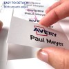 Изображение показывает руку, снимающую этикетку с листа, с именем «Paul Meyer» и логотипом «Avery», с примечанием, указывающим, что её легко отделить с гладкими краями.