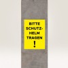 Изображение показывает желтый знак на бетонной колонне с надписью 'BITTE SCHUTZ-HELM TRAGEN!', что переводится как 'Пожалуйста, надевайте шлем!' указывая на требование безопасности.