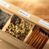 Изображение изображает деревянную коробку, содержащую различные чайные травы. Видны отсеки, помеченные различными видами чаев, включая ромашку (kamille) и цейлонский чай, с сушеными цветами ромашки, виднеющимися в одном отделении.