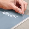 Рука снимает пластиковую обложку с тетради под названием «SCHULHEFT», что является немецким термином для «школьной тетради». На заднем фоне видна деревянная поверхность.