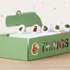 Изображение показывает зеленую коробку с наклейкой 'THINGS' и изображением головы динозавра сверху. Внутри находятся цветные карты, а на передней и верхней частях коробки наклеены несколько наклеек с ежиками.