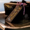 Изображение содержит декоративную подарочную бирку с надписью 'Frohe Weihnachten' (С Рождеством по-немецки), выполненной золотом. Бирка украшена различными праздничными символами, такими как звезды и санки, и свисает с упакованного подарка с красно-белой скрученной веревочкой. Обстановка отражает праздничную атмосферу с блестящей поверхностью.