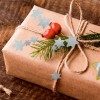 Изображение показывает красиво упакованный подарок в коричневую бумагу, украшенный бантом из натурального шпагата, зелеными листьями, красными ягодами и синими звёздочками.