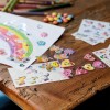Изображение представляет собой творческое рабочее пространство с красочными рисунками и различными стикерами. На бумаге нарисована радуга, украшенная блестящими камнями. На деревянном столе лежат несколько листов стикеров с мультяшными персонажами, сердцами и цветами, а также цветные карандаши и разбросанные конфеты.